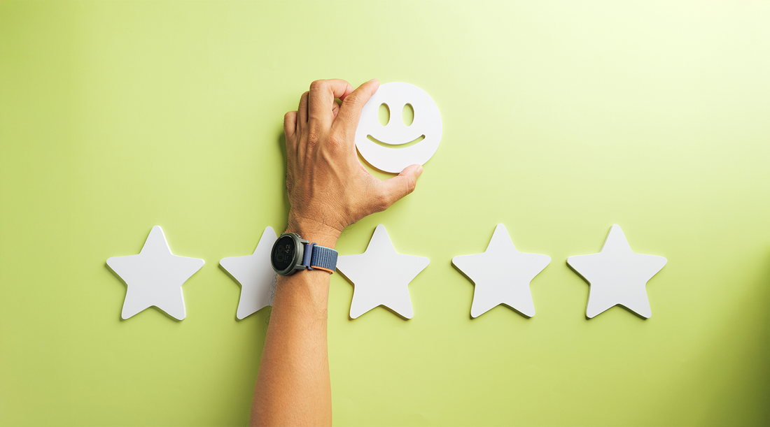 Uma mão humana posiciona um ícone circular de rosto sorridente (smiley) acima de uma fileira de cinco estrelas brancas em um fundo verde claro. A imagem simboliza excelência, satisfação do cliente e uma avaliação de pontuação máxima.