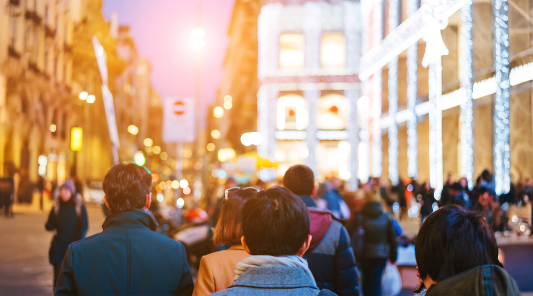 Multidão de pessoas vista de costas caminhando por uma rua urbana movimentada ao anoitecer. O fundo está desfocado e exibe prédios de arquitetura clássica com luzes quentes e fachadas iluminadas, criando uma atmosfera vibrante e cosmopolita.