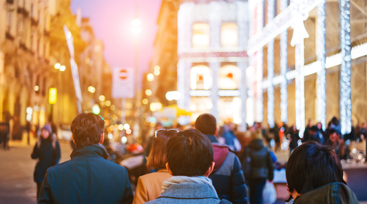 Multidão de pessoas vista de costas caminhando por uma rua urbana movimentada ao anoitecer. O fundo está desfocado e exibe prédios de arquitetura clássica com luzes quentes e fachadas iluminadas, criando uma atmosfera vibrante e cosmopolita.